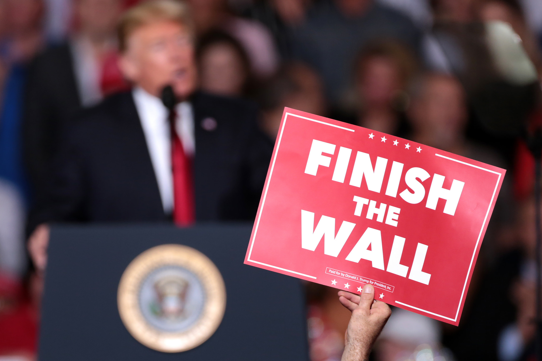 Donald Trump speaking at a rally, with a "finish the wall" sign in the foreground